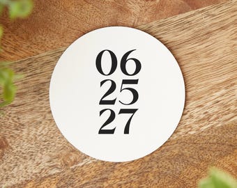 May include: A white, circular coaster with the numbers "06 25 27" printed in black. The coaster is set on a wooden surface with a natural grain pattern. Green foliage is visible in the background.