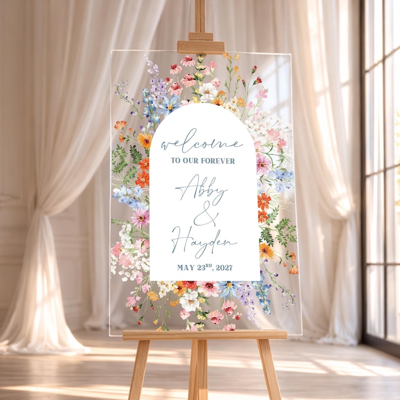 May include: A clear acrylic wedding welcome sign on a wooden easel. The sign features floral artwork in shades of pink, orange, blue, and white. Text includes "welcome to our forever", names, and a date.