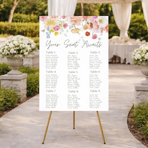May include: A white seating chart with the text "Your Seat Awaits" and table numbers 1-9. The top of the chart is decorated with a floral arrangement of pink, yellow, and orange flowers. The chart is displayed on a gold easel.