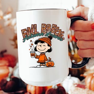 May include: White ceramic mug with a black rim and handle. The mug features a cartoon illustration of a girl holding a cup of coffee and a pumpkin. The text "Fall Breeze Autumn Leaves" is printed on the mug.