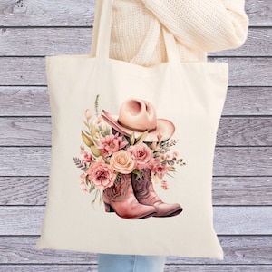 Könnte beinhalten: Eine weiße Canvas-Tragetasche mit einem rosafarbenen Cowboyhut und Stiefeln mit rosafarbenen Blumen. Die Tasche wird von einer Person gehalten.