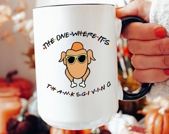 Può includere: Tazza in ceramica bianca con manico e bordo neri, con un tacchino dei cartoni animati con occhiali da sole. Il testo "THE ONE WHERE IT'S THANKSGIVING" è stampato sulla tazza con un carattere colorato. La tazza è tenuta da una persona.