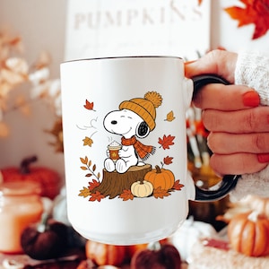 May include: A white ceramic mug featuring a graphic of Snoopy wearing an orange knit hat and scarf, sitting on a tree stump while holding a warm beverage. The design includes scattered autumn leaves and two pumpkins at the base of the stump.