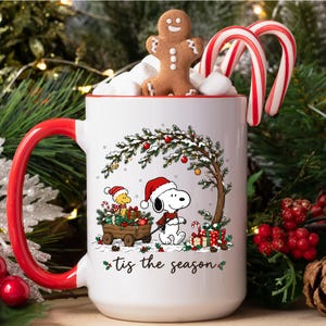 May include: A white ceramic mug with a red handle and rim, featuring a graphic of Snoopy and Woodstock with a wagon of gifts under a tree. The text 'tis the season' is printed below the illustration. The mug is filled with marshmallows, a gingerbread man, and candy canes.