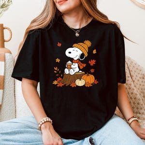 May include: A black t-shirt featuring a graphic of the cartoon character Snoopy wearing an orange knit hat and scarf, holding a coffee cup while sitting on a tree stump surrounded by orange and red autumn leaves and two orange pumpkins.