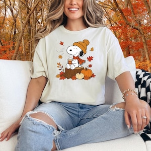 May include: A cream-colored short-sleeve t-shirt featuring a graphic of Snoopy wearing an orange knit hat and scarf, holding a steaming mug while sitting on a tree stump surrounded by orange pumpkins and falling autumn leaves.