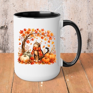 May include: A white ceramic mug with a black interior and handle, featuring a graphic of a tabby cat wearing a red and orange plaid scarf, sitting under an autumn tree with orange leaves, surrounded by several pumpkins on the ground.