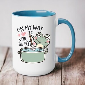 May include: White ceramic mug with a blue rim and handle. The mug features a cartoon frog stirring a pot with a wooden spoon. The text on the mug reads "On my way to stir the pot".