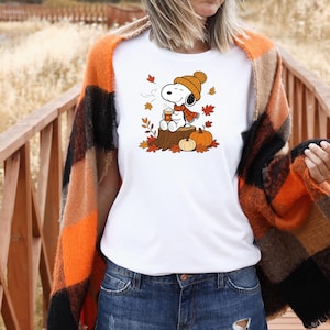 May include: A white t-shirt featuring a graphic of Snoopy wearing an orange knit hat and scarf, sitting on a tree stump while holding a coffee cup, surrounded by falling autumn leaves and two orange pumpkins.