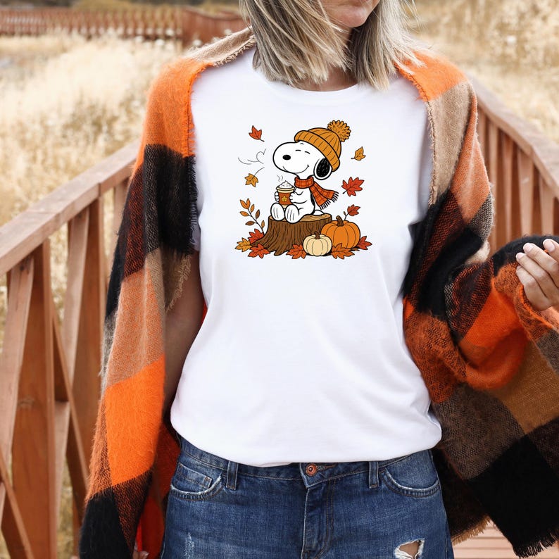 May include: A white t-shirt featuring a graphic of Snoopy wearing an orange knit hat and scarf, sitting on a tree stump while holding a coffee cup, surrounded by falling autumn leaves and two orange pumpkins.