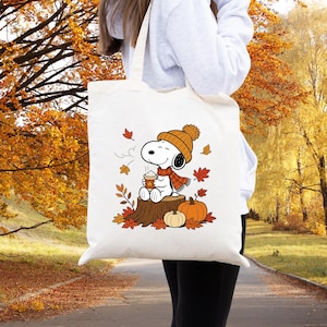 May include: A white canvas tote bag featuring a graphic of Snoopy wearing an orange knit hat and scarf, sitting on a tree stump while holding a steaming cup. The design includes falling autumn leaves and two pumpkins in shades of orange and cream.