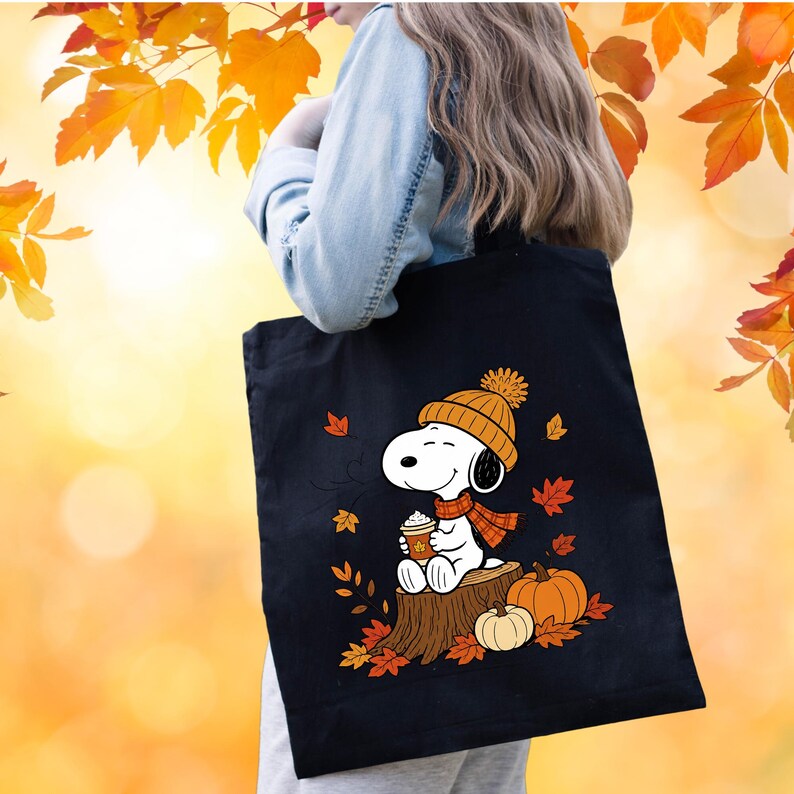 May include: A black tote bag featuring a graphic of the cartoon character Snoopy wearing an orange knit hat and scarf, sitting on a tree stump while holding a coffee cup, surrounded by orange and red autumn leaves and two orange pumpkins.