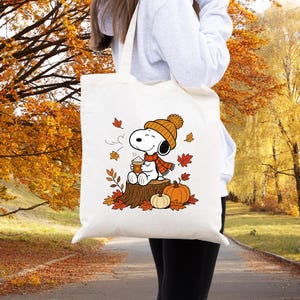 May include: A white canvas tote bag featuring a graphic print of a cartoon dog wearing an orange knit hat and scarf, sitting on a tree stump while holding a warm beverage, surrounded by orange pumpkins and scattered autumn leaves.