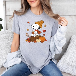 May include: A heather gray t-shirt featuring a graphic of Snoopy wearing an orange knit hat and scarf, sitting on a tree stump while holding a cup of coffee, surrounded by falling autumn leaves and two pumpkins on the ground.