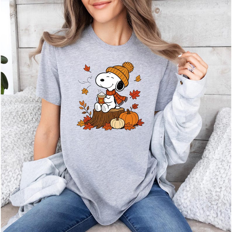 May include: A heather gray t-shirt featuring a graphic of Snoopy wearing an orange knit hat and scarf, sitting on a tree stump while holding a cup of coffee, surrounded by falling autumn leaves and two pumpkins on the ground.