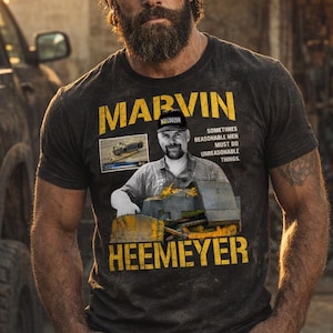 May include: Dark gray t-shirt with the name "MARVIN HEEMEYER" in yellow block letters. The shirt features a photo of a man in a bulldozer and the text "Sometimes Reasonable Men Must Do Unreasonable Things."