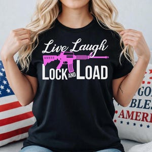May include: Black t-shirt with the words "Live Laugh Lock and Load" in white and a pink rifle graphic. The shirt has a crew neck and short sleeves. The text is in a cursive font.