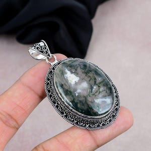 Puede incluir: Colgante ovalado con una piedra de ágata musgosa que muestra patrones verdes y blancos. La piedra está engastada en un marco decorativo de metal plateado con detalles intrincados. El colgante tiene una argolla para sujetar una cadena.