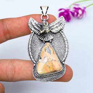 May include: A silver-toned pendant featuring a detailed eagle with outstretched wings positioned above a triangular, multi-colored stone with tan, cream, and grey veining. The metal frame has a textured, oxidized finish and a bail at the top.