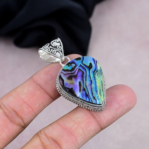 Könnte beinhalten: Herzförmiger Abalone-Muschel-Anhänger mit silberfarbener Öse. Der Anhänger zeigt eine Abalone-Muschel mit schillernden blauen, grünen und violetten Farbtönen. Die Muschel ist in einen dekorativen silberfarbenen Rahmen gefasst.