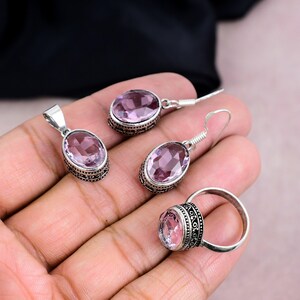 K&ouml;nnte beinhalten: Ein Schmuckset mit Anh&auml;nger, Ohrringen und einem Ring. Jedes St&uuml;ck hat einen ovalen, hellvioletten Edelstein, der in einem silberfarbenen Metall mit einem verzierten, dunklen Muster gefasst ist. Die Ohrringe sind als H&auml;ngeohrringe gestaltet.