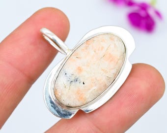 Può includere: Un ciondolo d'argento con una pietra ovale color crema. La pietra ha un aspetto maculato con sfumature di pesca e nero. Il ciondolo è tenuto tra due dita su uno sfondo bianco, con un fiore viola sfocato sullo sfondo.