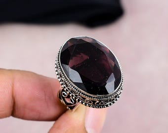 May include: A large, oval-shaped ring featuring a faceted, dark purple gemstone. The gemstone is set in a silver-colored band with intricate, ornate detailing around the stone and on the ring's shoulders. The ring is displayed against a soft pink background.