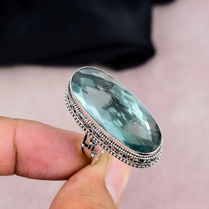 Può includere: Un anello ovale con una pietra preziosa azzurra. La fascia color argento presenta un motivo intricato attorno alla pietra. La pietra preziosa è incastonata in una lunetta. L'anello è tenuto da una mano.