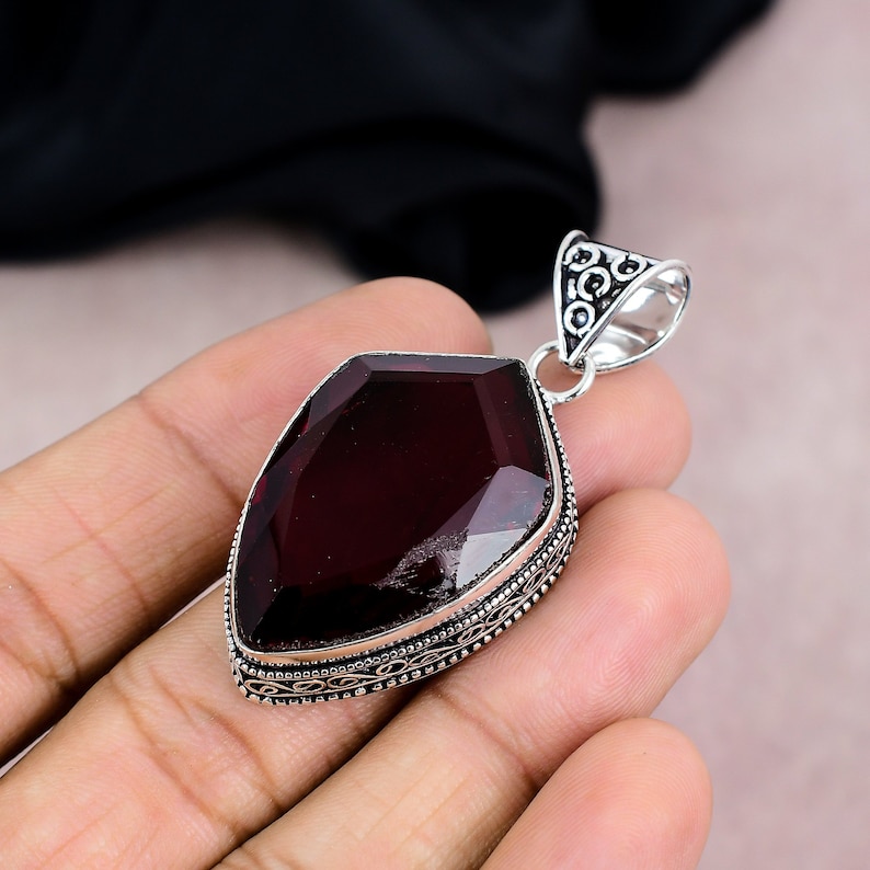 May include: A faceted, dark red gemstone pendant with a silver-colored bail. The pendant has an ornate, detailed border. The gemstone is a deep, rich color, and the bail has a decorative design.