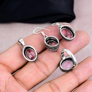 K&ouml;nnte beinhalten: Ein Schmuckset mit Anh&auml;nger, Ohrringen und einem Ring. Jedes St&uuml;ck hat einen ovalen, rosa Edelstein, der in einem silberfarbenen, verzierten Rahmen gefasst ist. Die Ohrringe haben einen Hakenverschluss.