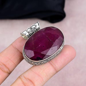Pode incluir: Um pingente grande, oval, com uma pedra preciosa vermelha escura facetada. A pedra preciosa está engastada numa moldura de metal prateado com uma argola decorativa. O pingente é segurado pelos dedos de uma pessoa.