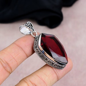 May include: A silver pendant with a large, faceted, rectangular, deep red gemstone. The pendant has an ornate, filigree-style border and a decorative bail. The gemstone is the focal point, catching the light and showcasing its rich color.