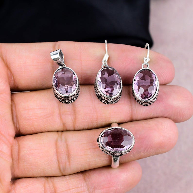K&ouml;nnte beinhalten: Ein Schmuckset mit Anh&auml;nger, Ohrringen und einem Ring. Jedes St&uuml;ck hat einen ovalen, hellvioletten Edelstein, der in einem silberfarbenen Metall mit einem verzierten, gemusterten Rand gefasst ist. Der Ring befindet sich an einem Finger.