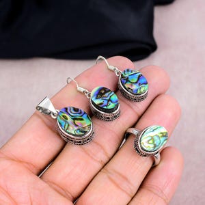 May include: Abalone shell jewelry set featuring a pendant, earrings, and ring. The oval-shaped abalone shells display iridescent blues, greens, and purples. The silver-toned metal settings have a detailed, patterned border.