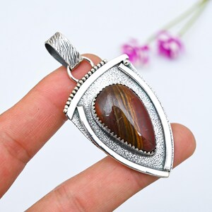 Puede incluir: Colgante de plata con forma de escudo. Presenta una piedra pulida de forma ovalada con rayas marrones y doradas. El engaste de plata tiene un borde texturizado y una fianza texturizada para una cadena.