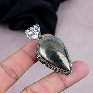 Puede incluir: Un colgante en forma de lágrima con una fianza de plata. El colgante presenta una piedra de pirita metálica pulida con un borde de cuentas de plata. La piedra tiene un color marrón dorado reflectante. El colgante es probablemente para un collar.