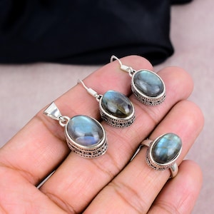 Puede incluir: Un conjunto de joyas que incluye un colgante, pendientes y un anillo. Cada pieza tiene una piedra ovalada, iridiscente azul-gris, engastada en un metal plateado con un borde ornamentado. El anillo está en un dedo.