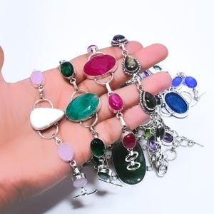 May include: A collection of gemstone necklaces with various shapes and colors. The necklaces feature oval, round, and teardrop-shaped stones in shades of green, pink, red, and blue, set in silver-toned metal. The gemstones are the focal point of the jewelry.