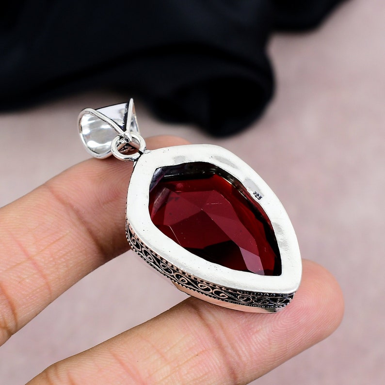 May include: A teardrop-shaped pendant with a faceted, deep red gemstone set in a silver metal frame. The pendant has an ornate, engraved border and a silver bail for a chain. The gemstone is the focal point, catching the light and showcasing its rich color.