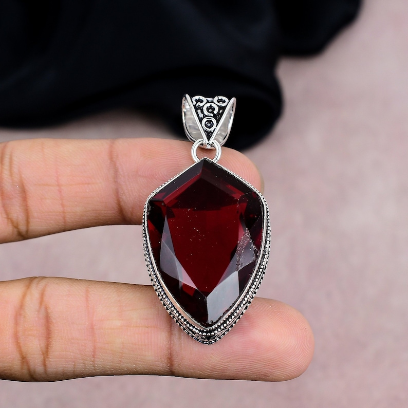 May include: A silver pendant with a large, faceted, deep red gemstone. The gemstone is shield-shaped and set in a detailed silver frame. The pendant has a decorative bail for a chain.