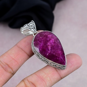 Pode incluir: Pingente em forma de gota com uma vibrante pedra preciosa magenta. O pingente tem uma moldura de prata detalhada com uma argola ornamentada. A superfície da pedra preciosa tem uma aparência texturizada.