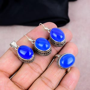 May include: A jewelry set featuring a pendant, earrings, and a ring. Each piece has a vibrant blue oval gemstone set in a silver-toned metal with an ornate border. The set is displayed on a hand.