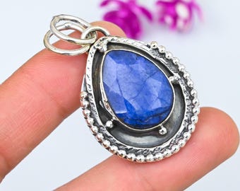 Op de afbeelding: Een druppelvormige hanger met een gefacetteerde, diepblauwe edelsteen, gezet in een zilverkleurig metalen frame. De rand van het frame is voorzien van kralen en een lus voor een ketting. De hanger wordt door een vinger tegen een witte achtergrond gehouden.