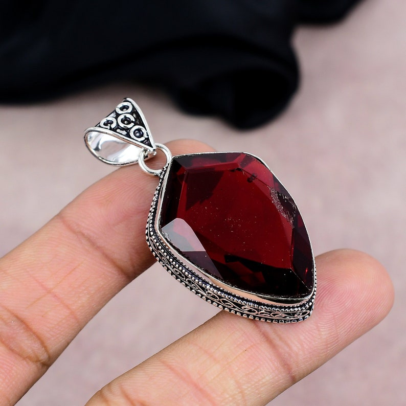 May include: A silver pendant featuring a large, faceted, deep red gemstone. The gemstone is set in a decorative silver frame with intricate detailing. The pendant has a bail for attaching to a chain.