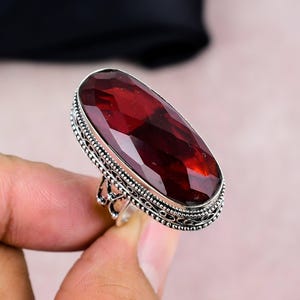 May include: A silver ring featuring a large, faceted, oval-shaped, deep red gemstone. The ring has an ornate, filigree-style setting around the gemstone and on the band. The gemstone is the focal point of this jewelry piece.