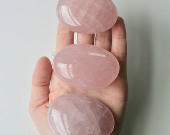 May include: Three smooth, pink rose quartz stones, also known as love stones, are stacked on top of each other in a person's hand. The stones are polished and have a natural, cloudy appearance.