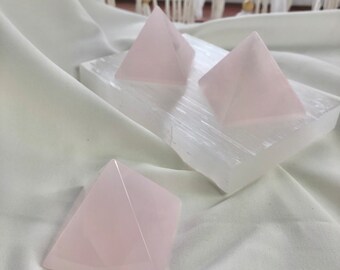 Op de afbeelding: Drie roze rozenkwarts kristallen piramides op een witte seleniet lader. De piramides zijn van verschillende grootte en zijn in een driehoekige vorm gerangschikt.