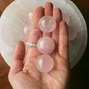 May include: A collection of pink rose quartz spheres, some of which are being held in a hand. The spheres are smooth and translucent, with a slight pink hue.