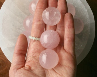 May include: A collection of pink rose quartz spheres, some of which are being held in a hand. The spheres are smooth and translucent, with a slight pink hue.