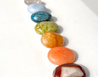 May include: Seven smooth, polished oval-shaped stones in a rainbow of colors, including clear, light blue, teal, green, yellow, orange, and red.